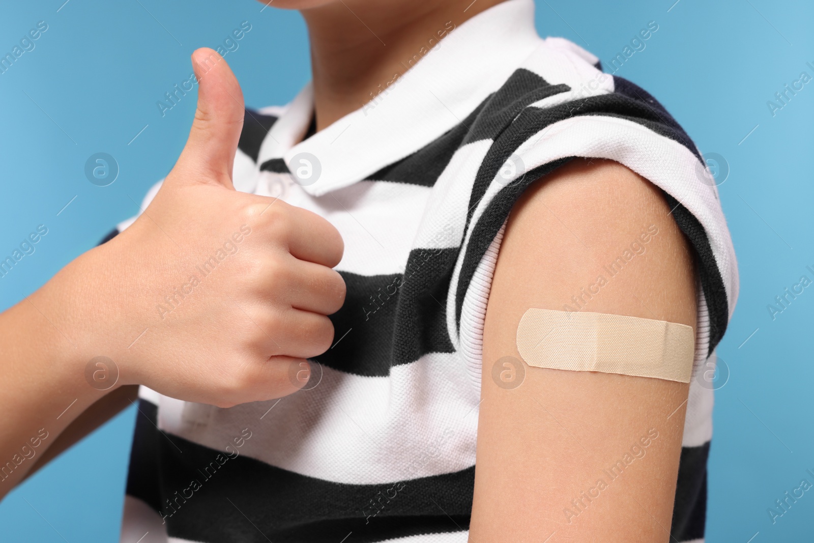 Photo of Boy with sticking plaster on arm after vaccination showing thumbs up against light blue background, closeup