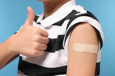 Boy with sticking plaster on arm after vaccination showing thumbs up against light blue background, closeup