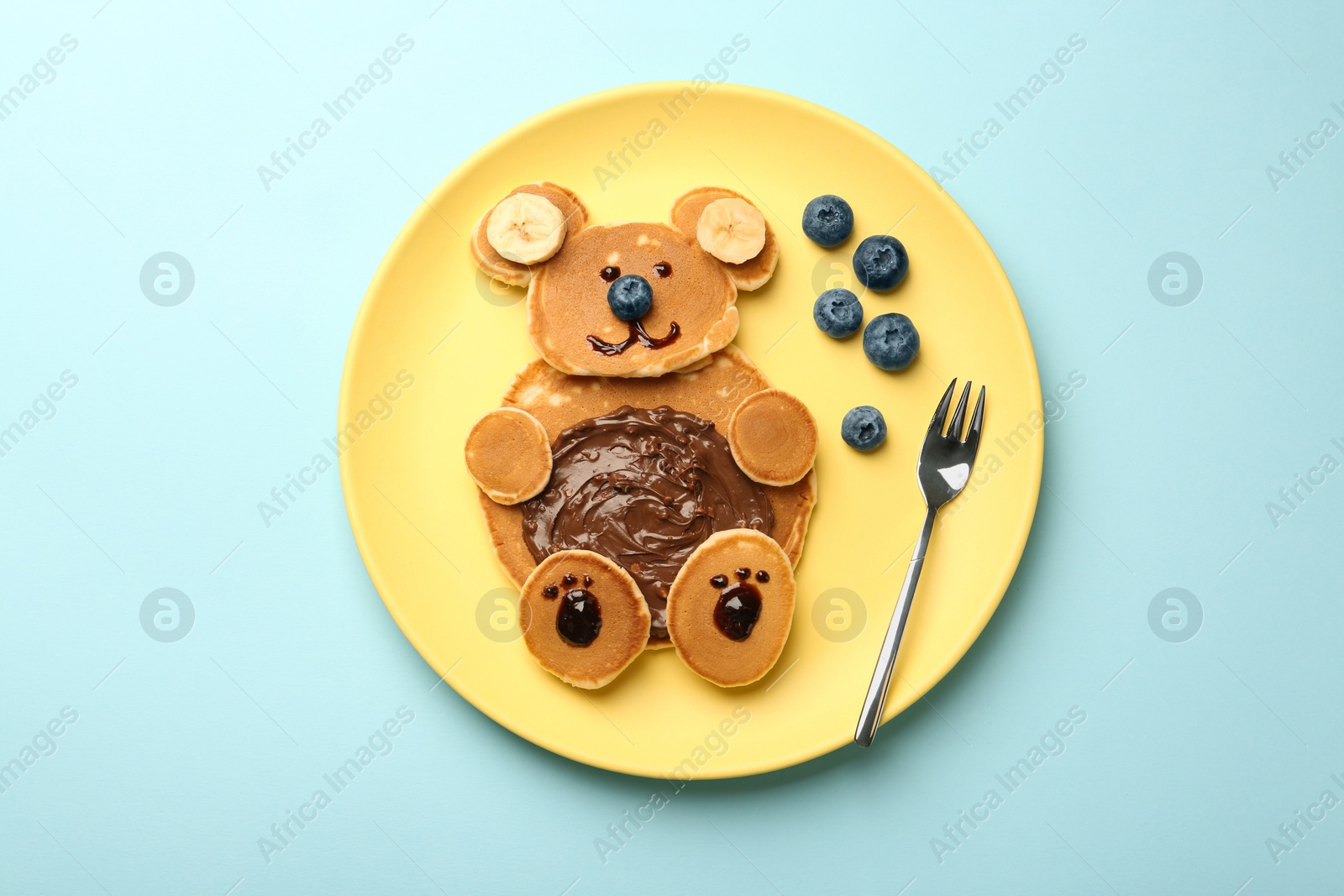 Photo of Creative serving for kids. Plate with cute bear made of pancakes, blueberries, bananas and chocolate paste on light blue table, top view