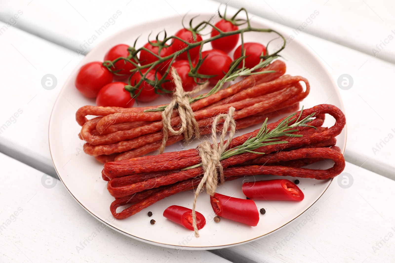 Photo of Bundles of delicious kabanosy with rosemary, peppercorn, chilli and tomatoes on white wooden table