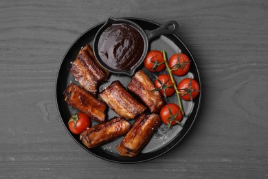 Photo of Tasty roasted pork ribs served with sauce and tomatoes on grey wooden table, top view