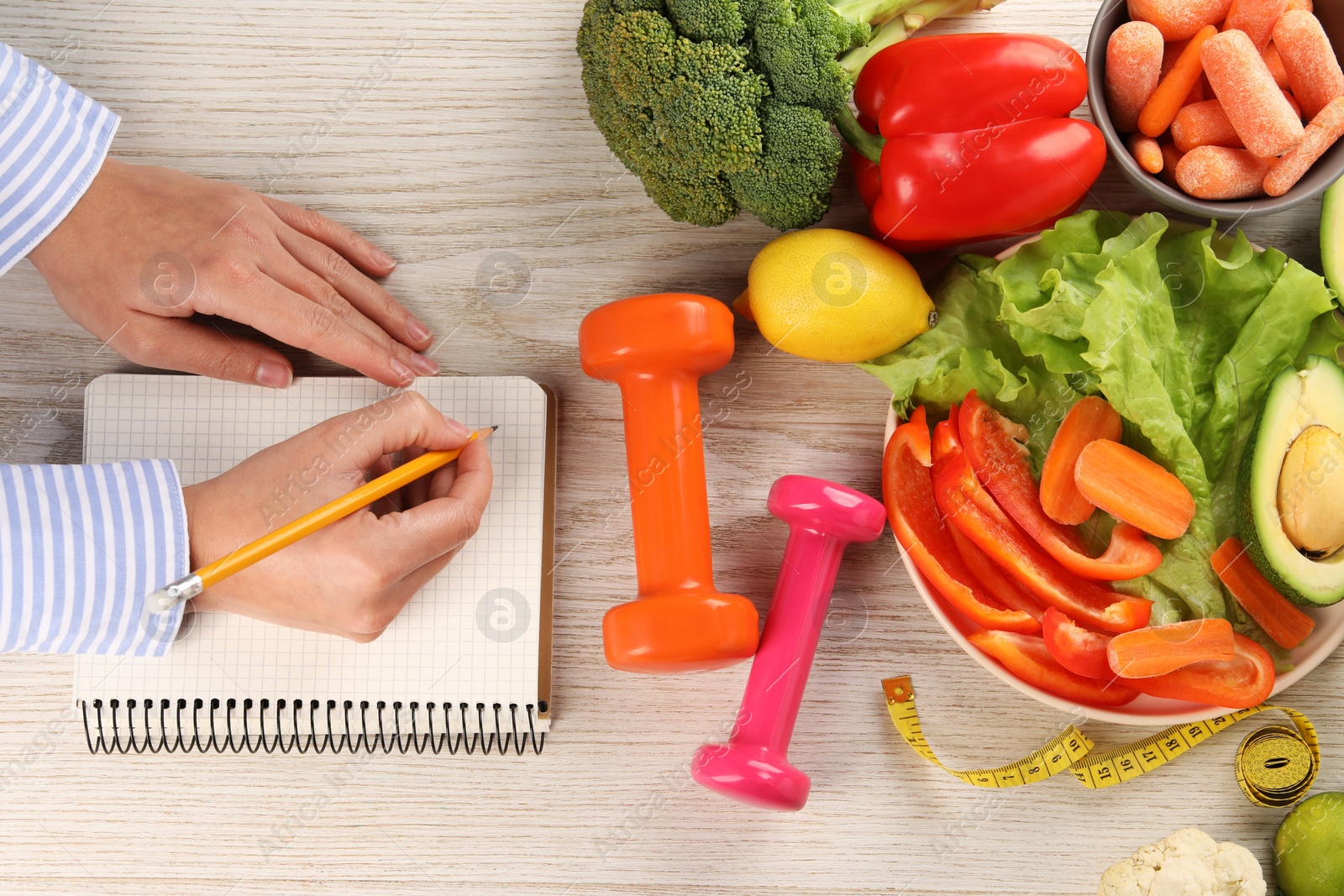 Photo of Woman developing diet plan at light wooden table with vegetables, dumbbells and measuring tape, top view