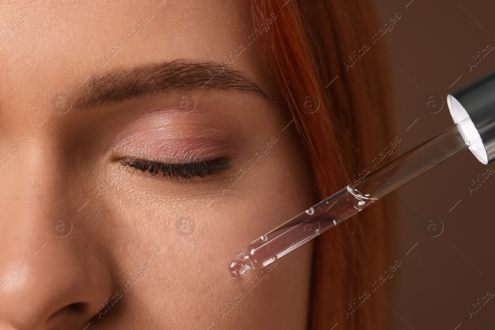 Photo of Beautiful young woman applying cosmetic serum onto her face on brown background, closeup