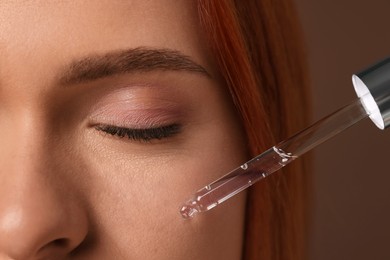 Photo of Beautiful young woman applying cosmetic serum onto her face on brown background, closeup