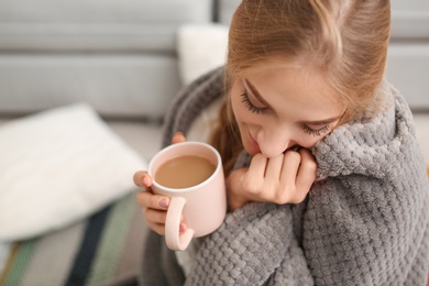 Beautiful young woman wrapped in plaid sitting with cup of coffee on floor at home. Winter atmosphere