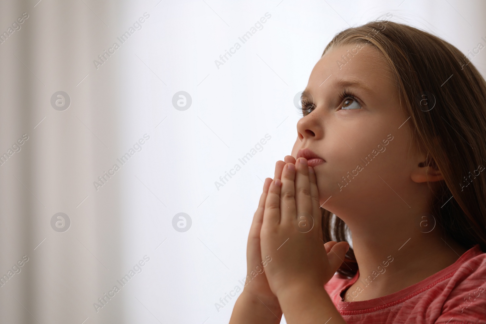 Photo of Cute little girl with hands clasped together praying on blurred background. Space for text