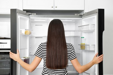 Photo of Young woman near empty refrigerator in kitchen, back view