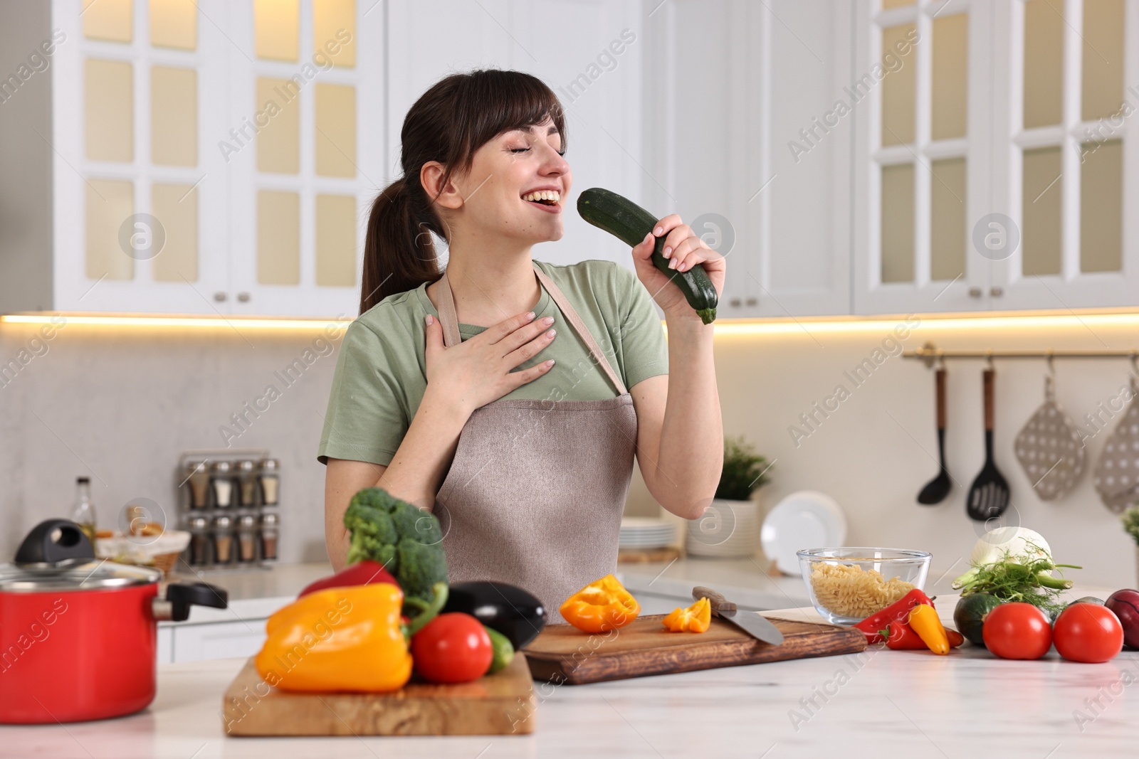 Photo of Happy young housewife with fresh cucumber having fun while cooking at white marble table in kitchen