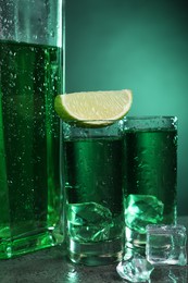 Photo of Absinthe in shot glasses with lime and ice cubes on gray textured table against green background, closeup. Alcoholic drink