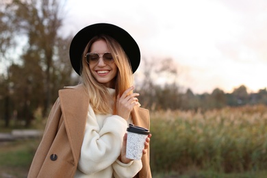 Photo of Beautiful young woman with cup of coffee wearing stylish autumn clothes outdoors