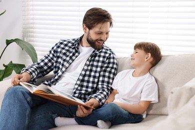 Photo of Happy dad and son reading book together on sofa at home