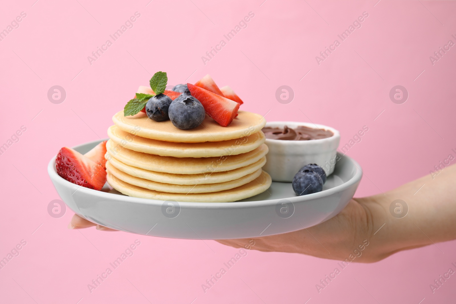 Photo of Woman holding delicious pancakes with strawberries, blueberries and chocolate sauce against pink background, closeup