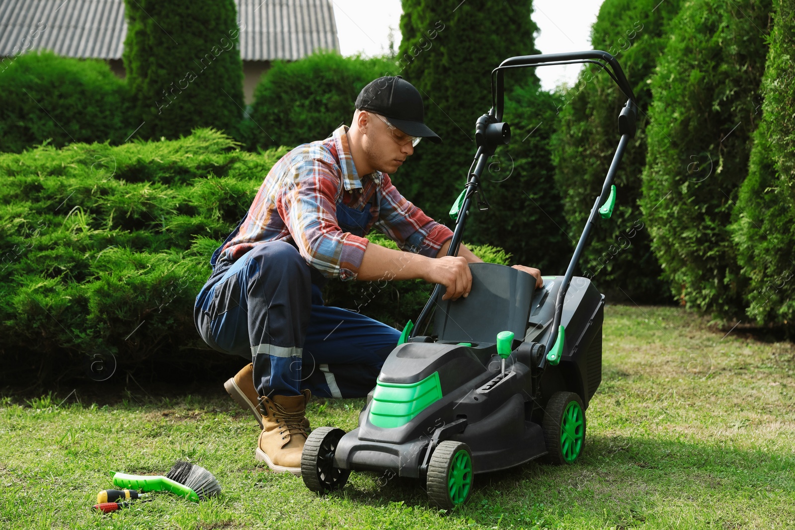 Photo of Cleaning lawn mower. Young man detaching grass catcher from device in garden