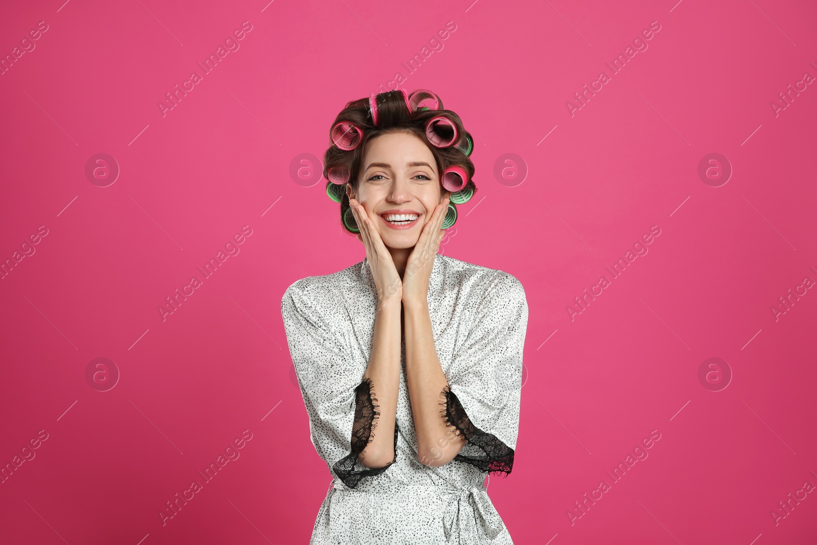 Photo of Beautiful young woman in silk bathrobe with hair curlers on pink background