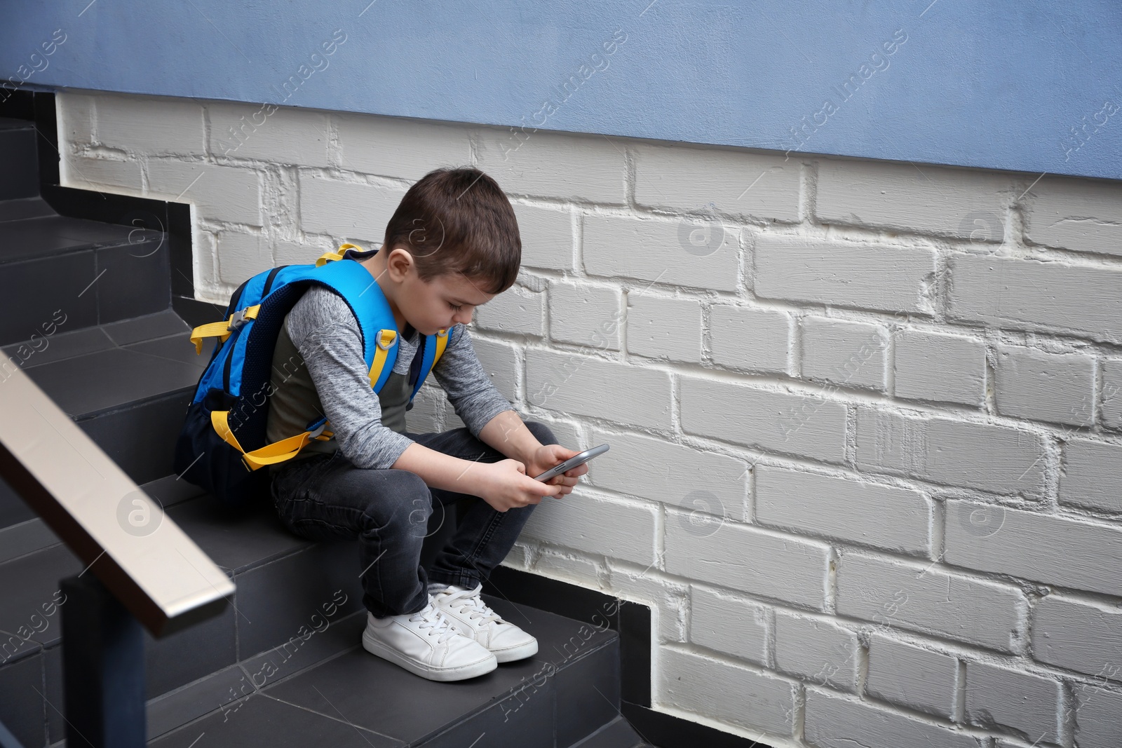 Photo of Sad little boy with mobile phone sitting on stairs indoors