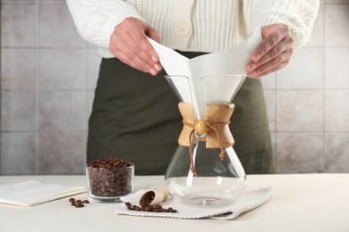 Photo of Making drip coffee. Woman setting paper filter into glass chemex coffeemaker at table, closeup