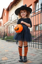 Photo of Cute little girl with pumpkin candy bucket wearing Halloween costume outdoors