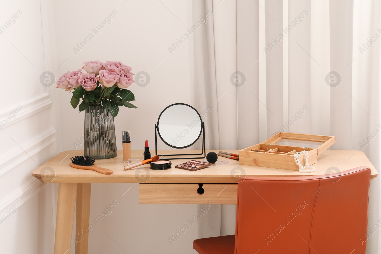 Photo of Mirror, cosmetic products, box of jewelry and vase with pink roses on wooden dressing table in makeup room