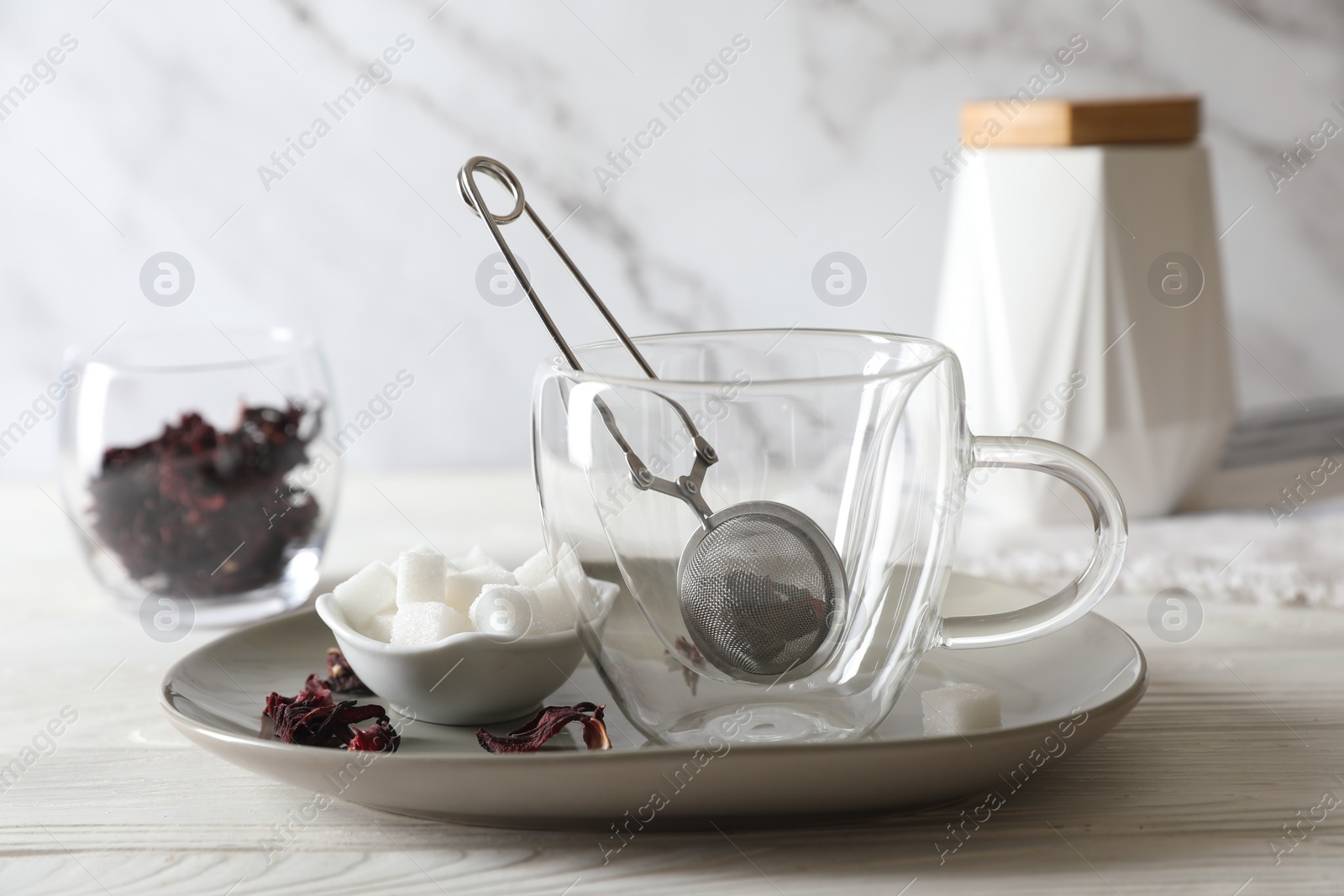 Photo of Making hibiscus tea. Cup with infuser, sugar cubes and dried roselle petals on white wooden table