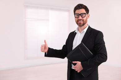 Photo of Happy real estate agent with leather portfolio showing thumb up in new apartment. Space for text