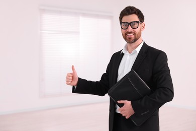 Happy real estate agent with leather portfolio showing thumb up in new apartment. Space for text