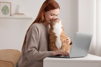 Photo of Woman working with laptop and hugging cat at desk. Home office