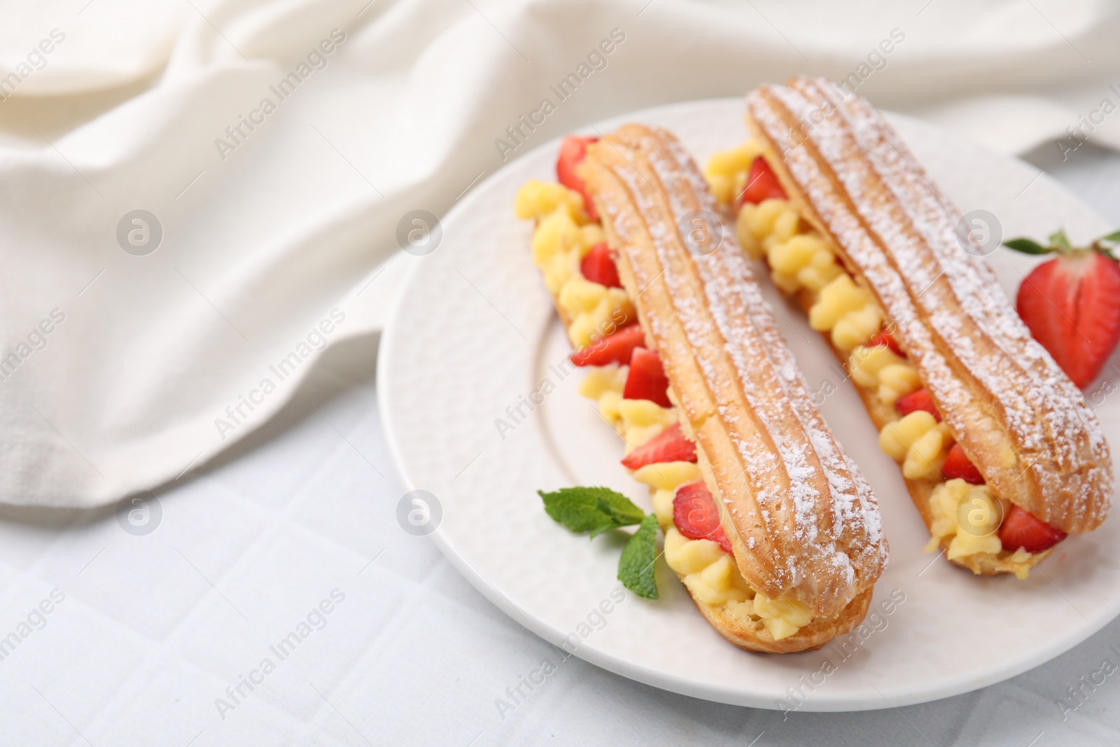 Photo of Delicious eclairs filled with cream, strawberries and mint on white tiled table, closeup