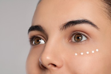 Photo of Young woman with cream on skin under eye against light grey background, closeup
