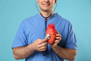 Doctor with stethoscope and model of heart on light blue background, closeup. Cardiology concept