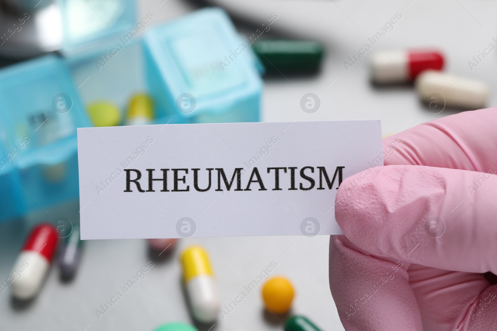 Photo of Doctor in glove holding sheet of paper with word Rheumatism above table with pills, closeup