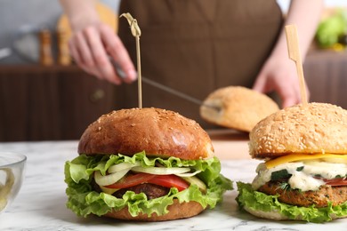 Photo of Woman cutting bun at white marble table, focus on delicious vegetarian burgers