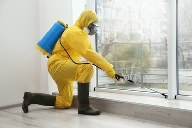 Photo of Pest control worker in protective suit spraying pesticide near window indoors