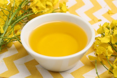 Photo of Rapeseed oil in bowl and beautiful yellow flowers on table, closeup