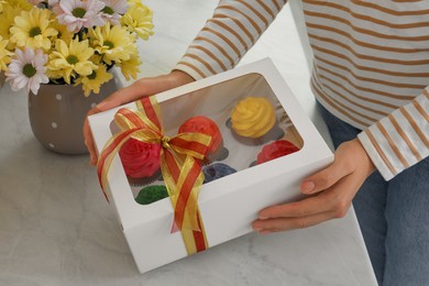 Photo of Woman holding box with delicious colorful cupcakes at white table, closeup