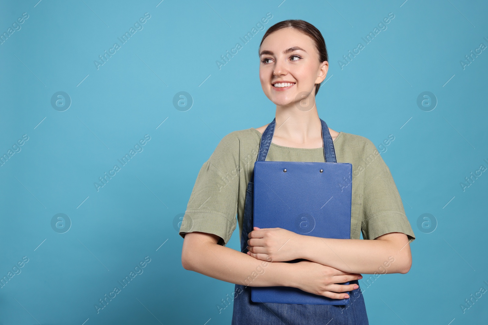 Photo of Beautiful young woman in clean denim apron with clipboard on light blue background. Space for text
