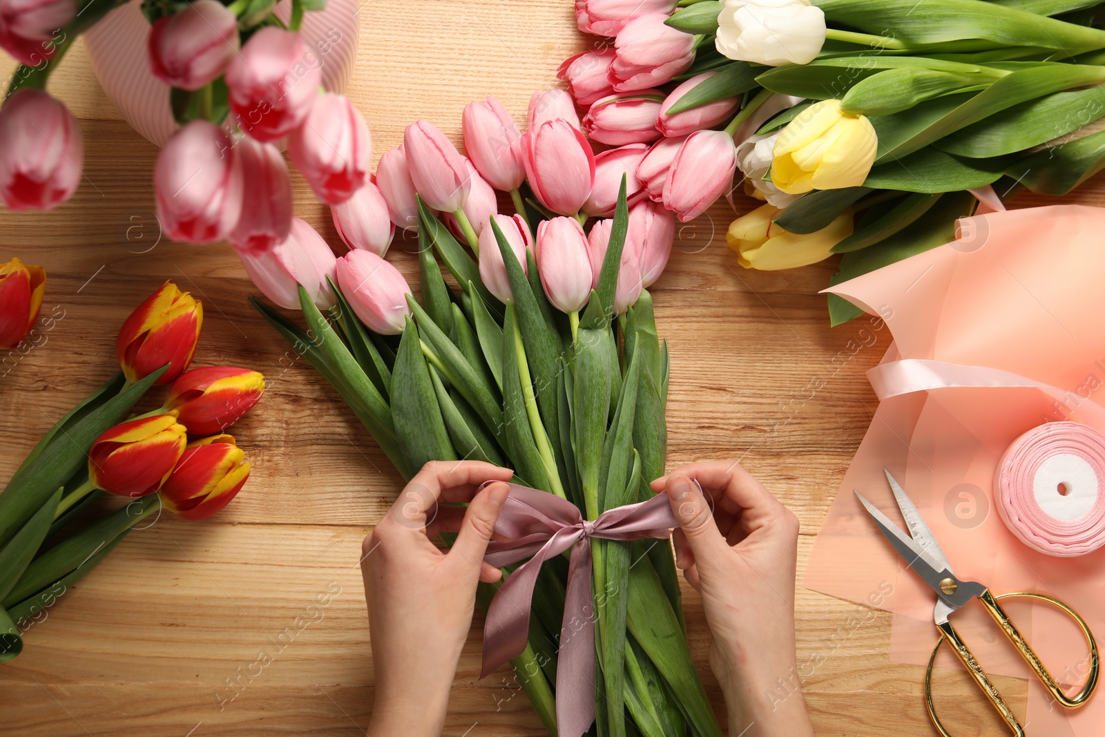 Photo of Woman making beautiful bouquet of fresh tulips and ribbon at wooden table, top view