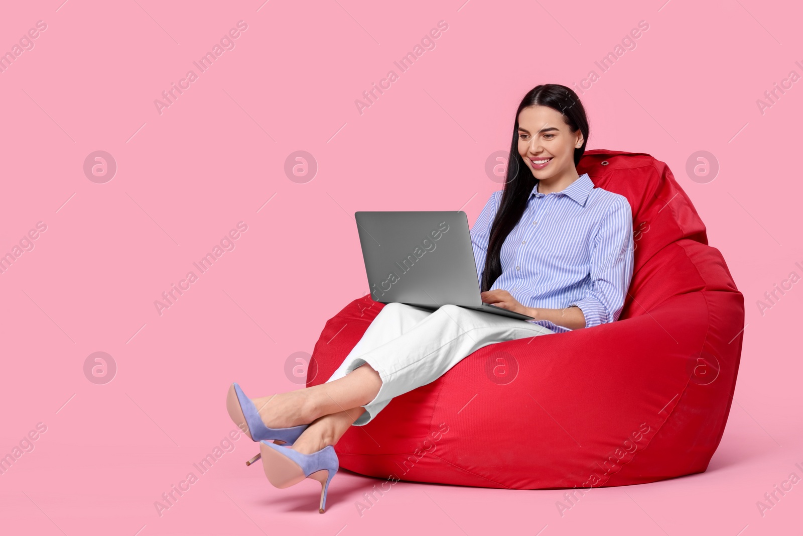 Photo of Happy woman with laptop sitting on beanbag chair against pink background