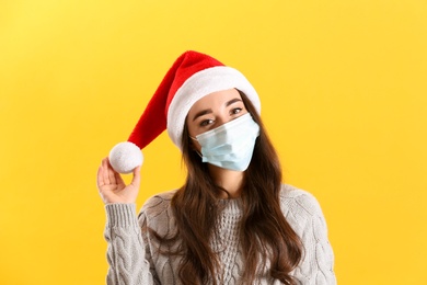 Photo of Beautiful woman wearing Santa Claus hat and medical mask on yellow background