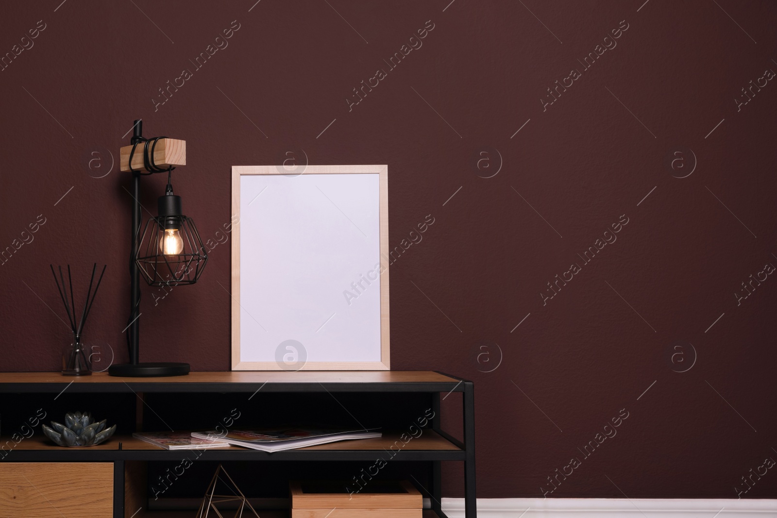 Photo of Empty frame and stylish lamp on wooden table near brown wall. Mockup for design