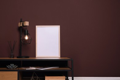 Empty frame and stylish lamp on wooden table near brown wall. Mockup for design