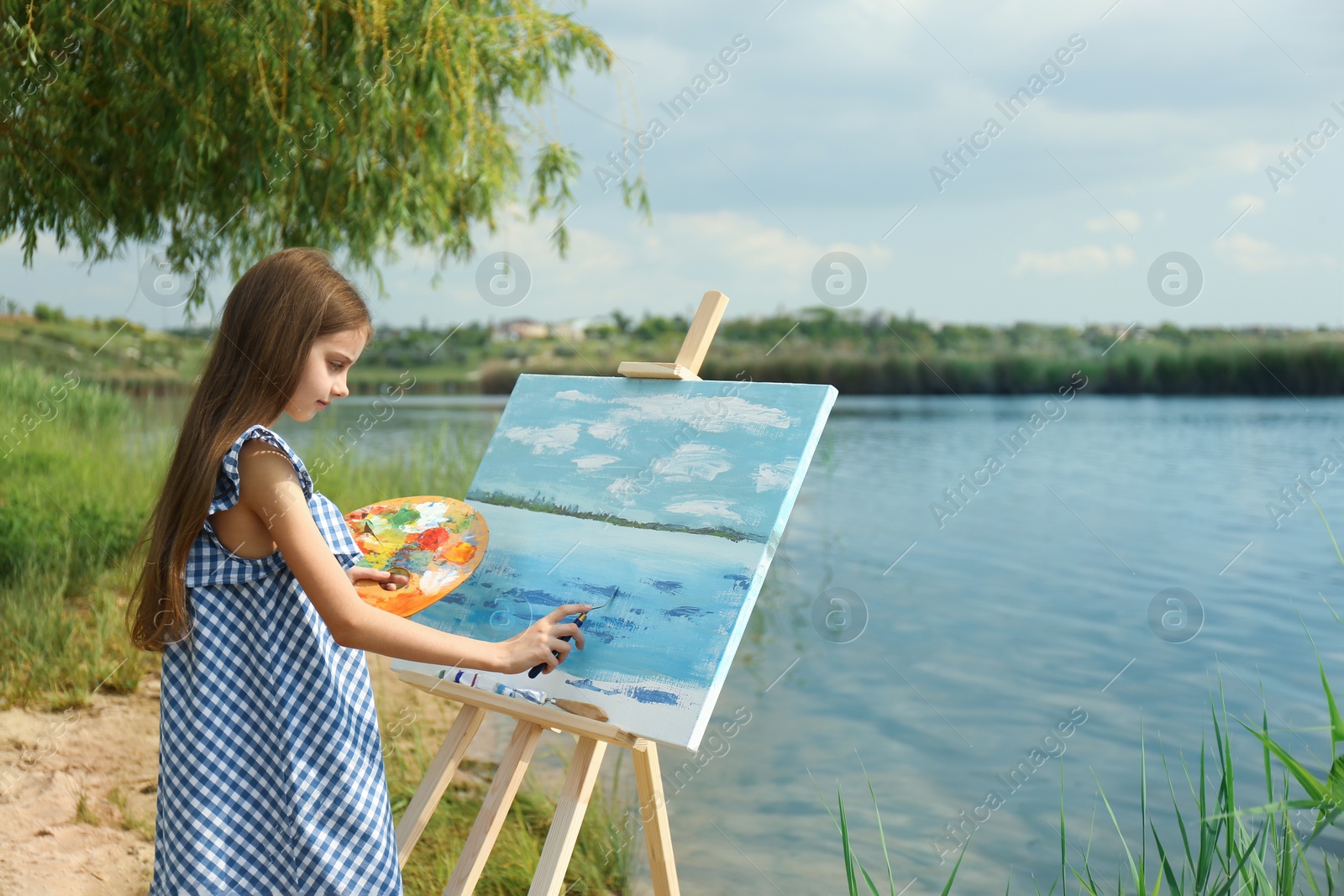 Photo of Little girl painting scenery on easel near lake