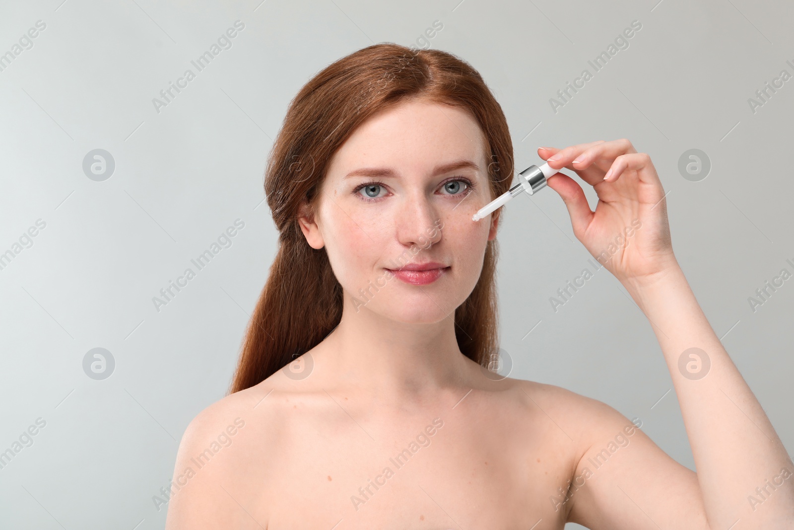 Photo of Beautiful woman with freckles applying cosmetic serum onto her face against grey background