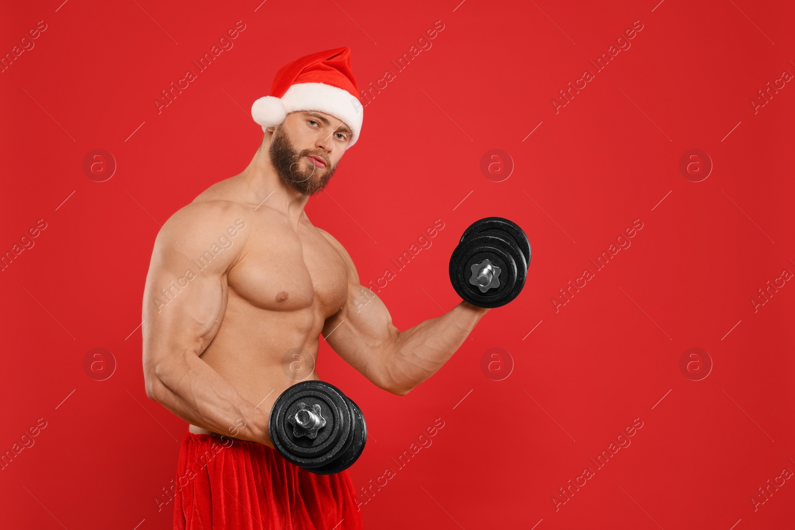 Photo of Muscular young man in Santa hat with dumbbells on red background, space for text