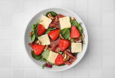 Photo of Tasty salad with brie cheese, prosciutto, strawberries and walnuts on white tiled table, top view