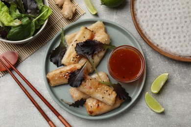 Photo of Tasty fried spring rolls, lettuce, lime and sauce on grey table, flat lay