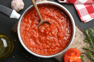 Photo of Homemade tomato sauce in pot, spoon and fresh ingredients on dark table, flat lay