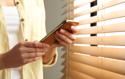 Young woman using modern smartphone near window indoors, closeup