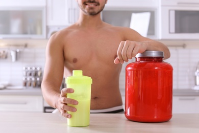 Photo of Young shirtless man holding protein shake in bottle at table with jar of powder at home