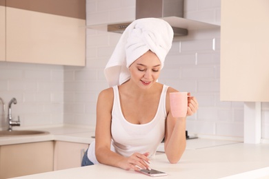 Beautiful woman with towel on head drinking tea and using smartphone in kitchen