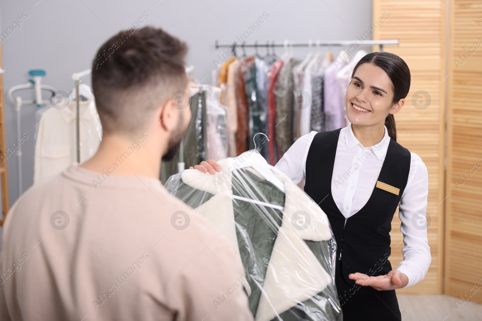 Photo of Dry-cleaning service. Happy worker giving coat in plastic bag to client indoors